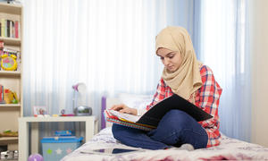 Young student in head wrap reading materials in their lap seated on a bed.