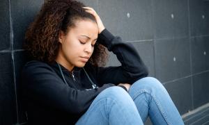 Young person of color in visible distress sitting with their back against a wall, hand on their head.