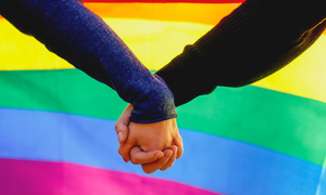 Two people hold hands with a rainbow flag in the background. 