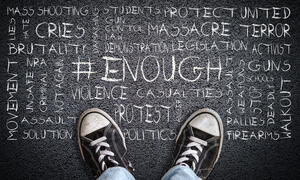 A child's feet with sneakers on them stand in front of words that include protect, students, mass shooting, cries, brutality, activism, walkout and more etched in chalk on the ground.