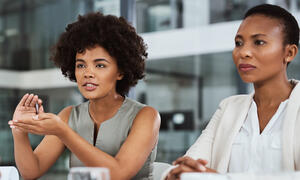 Two women sit at a table and one of the women is talking to someone across from her who is not in view. They are in a workshop setting.