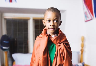 A kid standing on the bed with and adult shirt as a cape