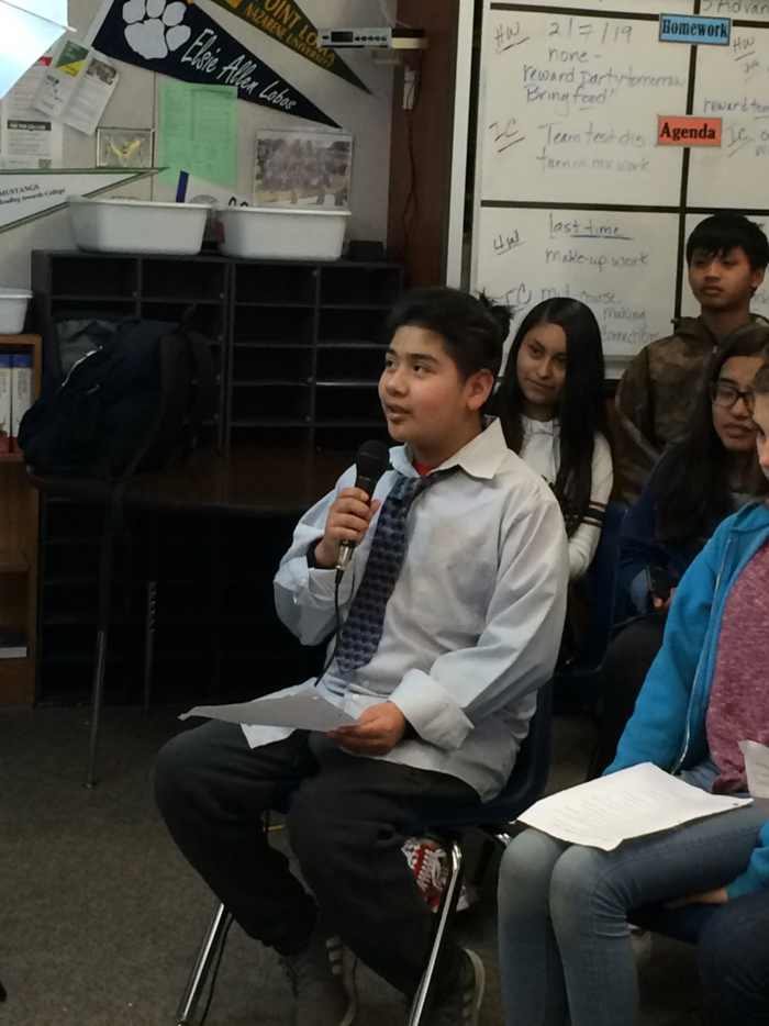 Young student wearing a tie holding a microphone asking a question of the interviewee, who is offscreen.